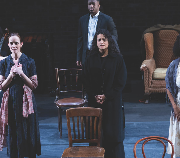 A group of actors and a cellist perform on stage, evoking deep emotions with their expressions. Three actors stand in the foreground, each expressing intense feelings through their posture and facial expressions, while another actor and a cellist are positioned in the background. The set includes various chairs and an armchair, creating a minimalist yet poignant scene.