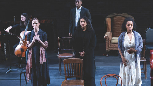 A group of actors and a cellist perform on stage, evoking deep emotions with their expressions. Three actors stand in the foreground, each expressing intense feelings through their posture and facial expressions, while another actor and a cellist are positioned in the background. The set includes various chairs and an armchair, creating a minimalist yet poignant scene.