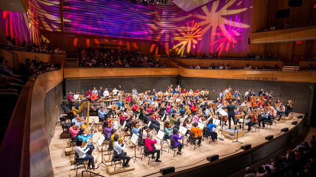 The photo captures a vibrant orchestral performance in progress inside a modern concert hall. The musicians are seated in a semi-circular arrangement on stage, surrounded by wood-paneled walls that reflect the colorful, abstract light patterns projected above them, creating a dynamic and festive atmosphere. A conductor is visible at the forefront, leading the orchestra. The audience, seated in the curved balconies and the main floor, is attentively watching the performance in a well-lit, packed venue.