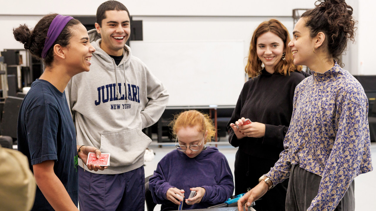 A group of vibrant Juilliard students in a workshop setting, laughing and engaging with each other as some of them hold playing cards, practicing their magic skills. The environment is casual and full of positive energy, with a sense of camaraderie and shared excitement for what they are learning.