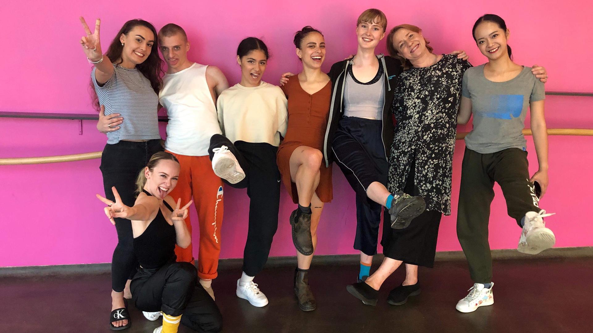 Juilliard dancers pose for a photo with dancers from the Trinity Laban Conservatory of Dance in London--names unknown--in the dance studio. They are wearing street clothes and behind them is a distinctive pink wall.