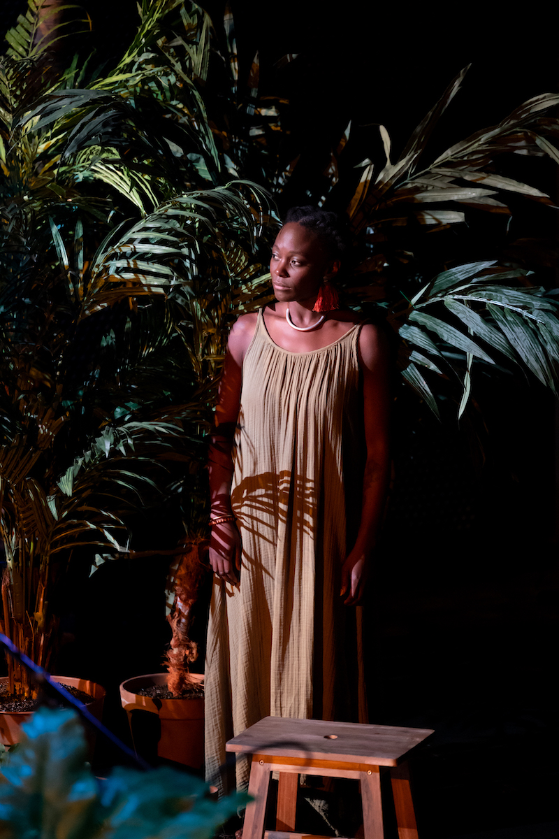 Nathalie Joachim in the midst of performance, making an intense facial expression, and she is surrounded by plants