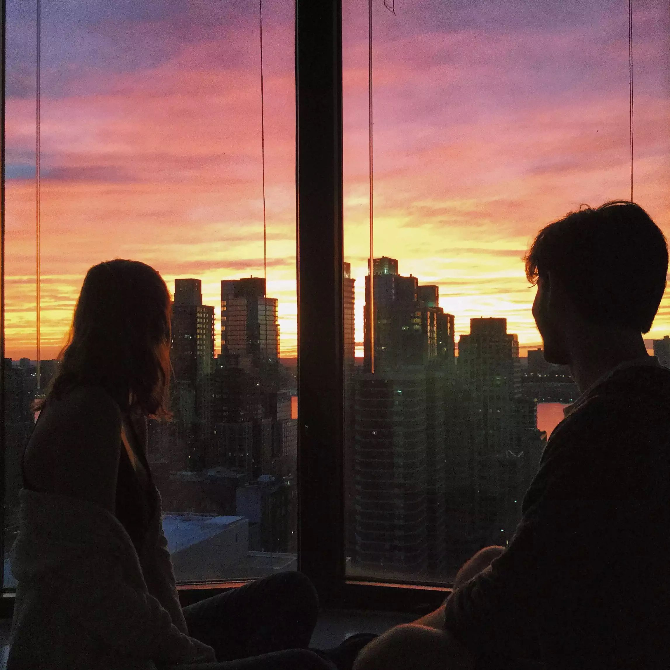 Two people sit by a window overlooking the skyline at sunset