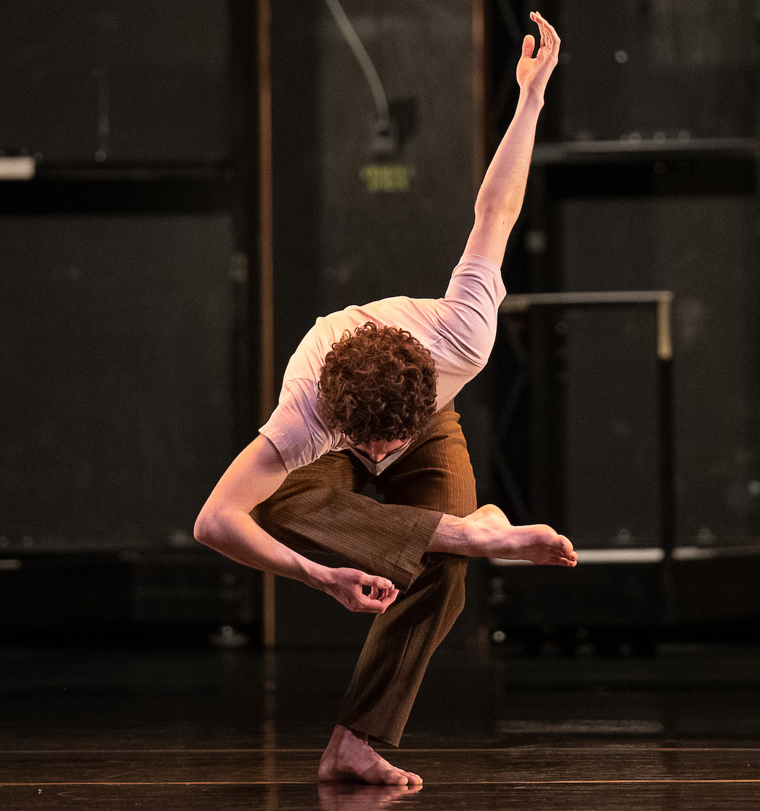 A dancer on one leg, the other crossed in front of him, making a deep bow in a strong and triumphant pose with his left arm pointing up 