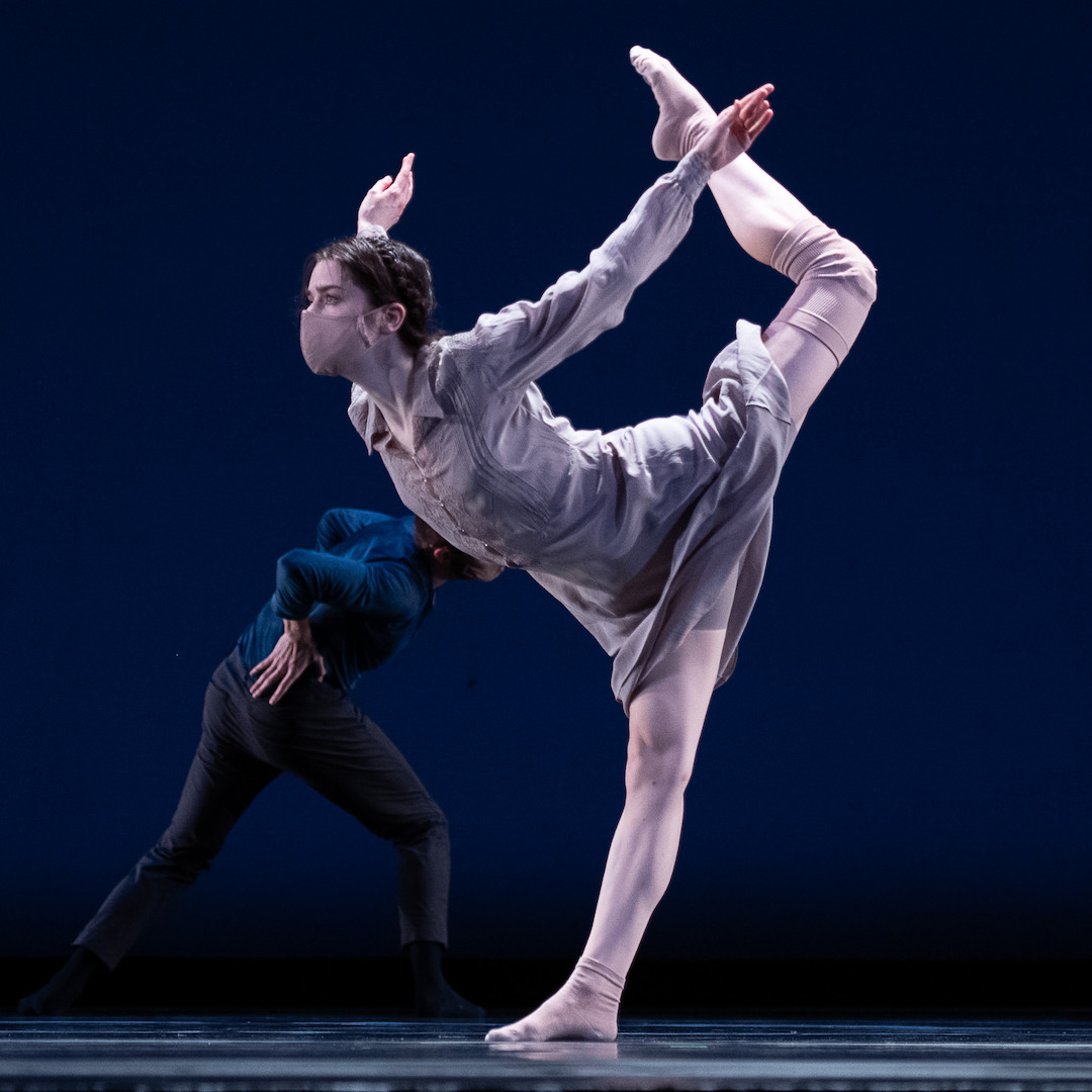 A dancer on one leg, costumed in silver, bends forward with arms extended behind her like a "V" as though flying