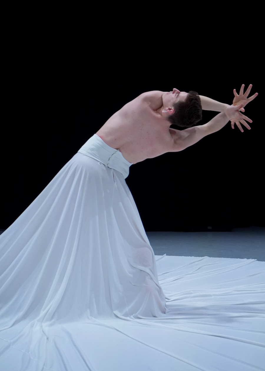 Dancer in a dramatic pose, leaning backward with arms extended and hands intertwined. The dancer is wearing a long, flowing white skirt that spreads out around them, creating a striking contrast against the dark background