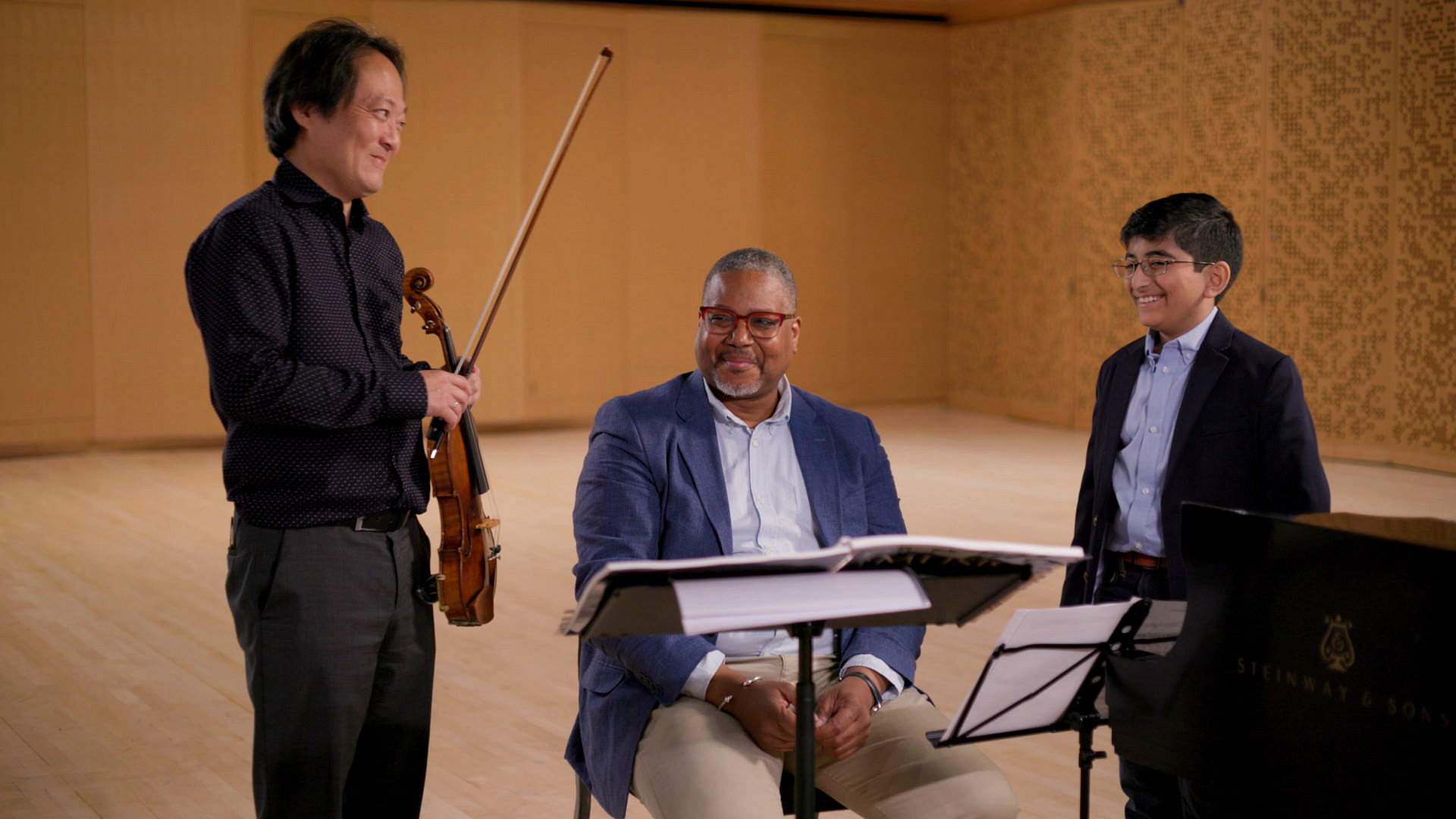 The three individuals captioned reviewing sheet music in a rehearsal studio. All are smiling and Yoo holds a violin.