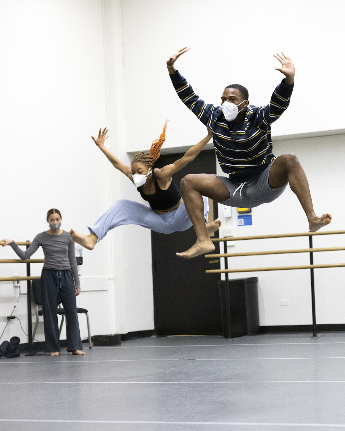 Two dancers are in mid-air while a third dancer looks on during rehearsal in a dance studio