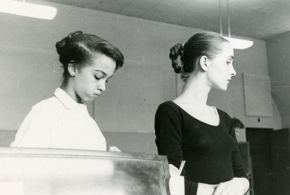 The archival (black and white) image depicts two young women engaged in a dance class. The woman on the left, Mercedes Ellington, is wearing a white blouse with her hair styled up, looking down at something she is holding. The woman on the right, Pina Bausch, is dressed in a dark ballet top, her hair pulled back into a neat bun, and is gazing with a focused expression. They both exhibit a posture of concentration and practice, suggesting a moment of learning or refinement of their craft. In the background, partially obscured, there is someone who might be a teacher or another student, providing context that this is an educational setting.