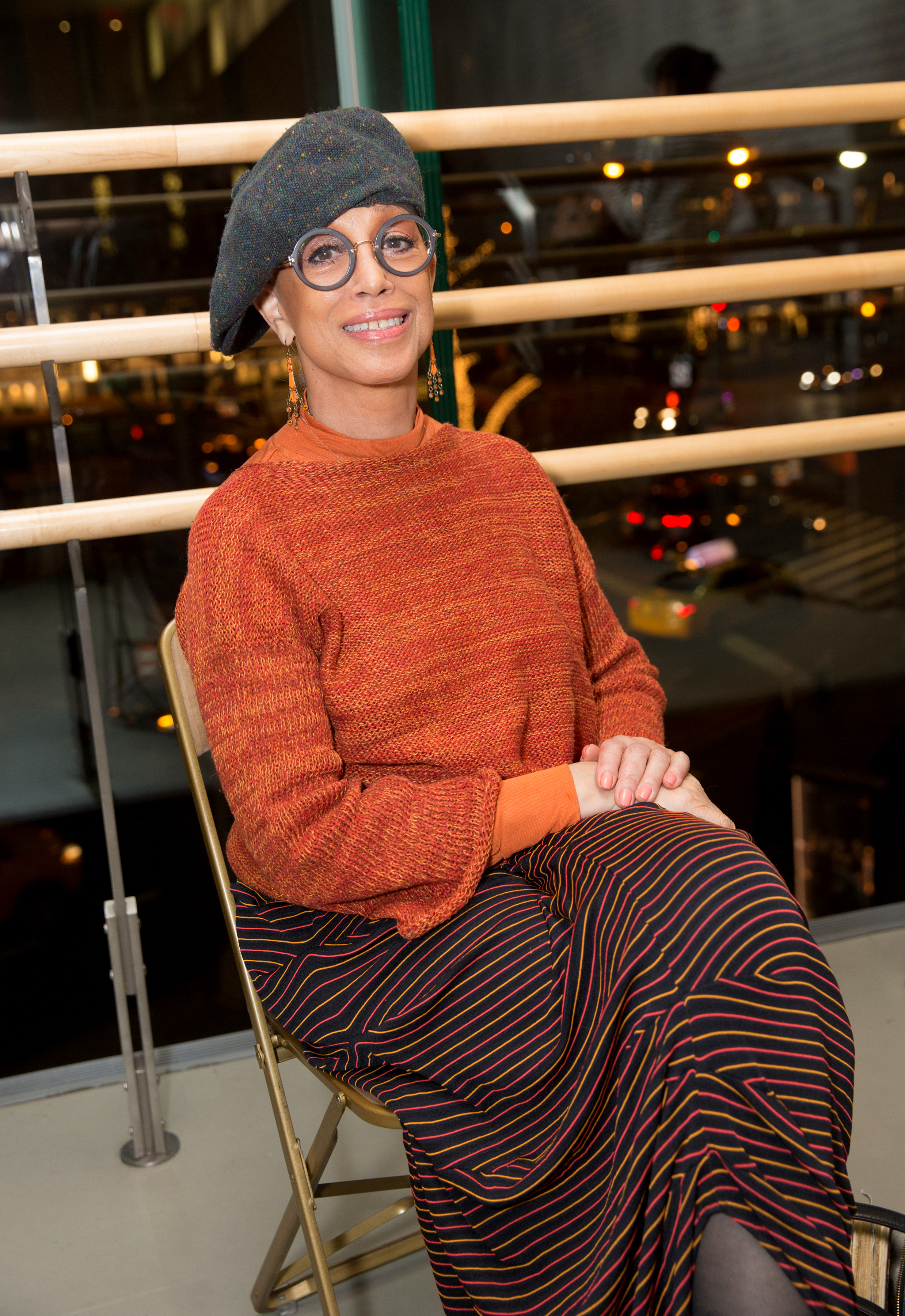 The image features a woman seated on a metal folding chair, smiling towards the camera. She's stylishly attired in a rust-colored knitted sweater and a beret, with large circular earrings complementing her look. Her outfit is completed with boldly patterned pants that include stripes and swirls in contrasting colors. 