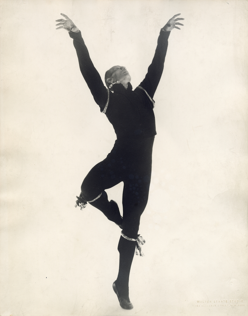 Archival (black and white) photo of José Limón performing. His arms are extended up and he is standing on one leg. His costume covers almost his entire body, leaving only his hands and head exposed.
