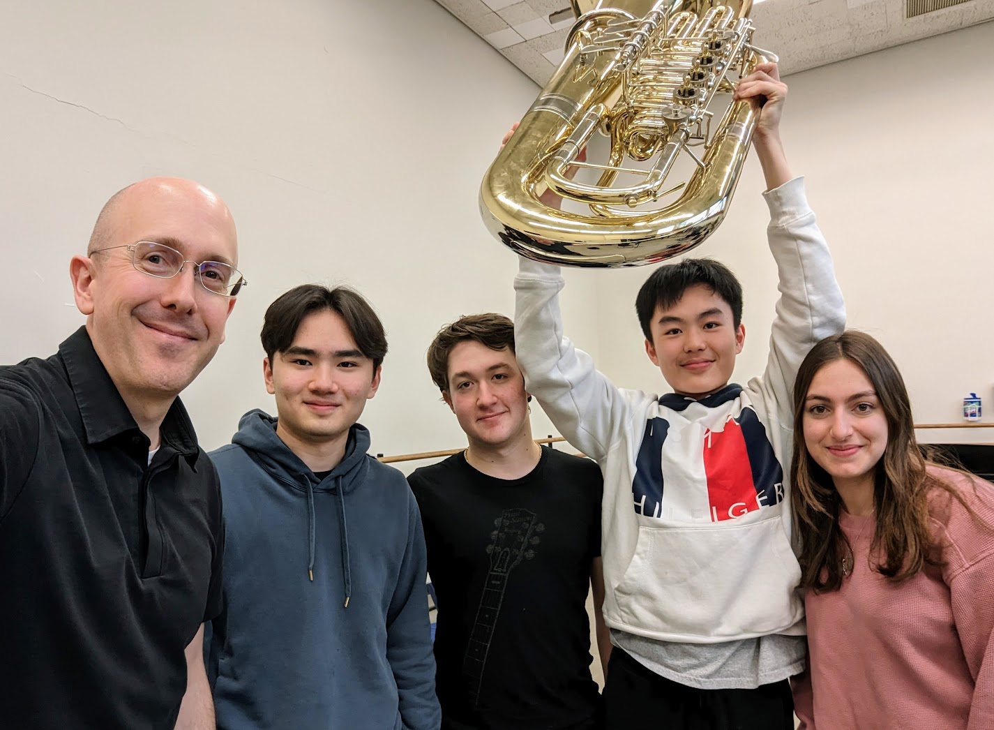 The students and their teacher stand in a semi-circle. They are dressed casually and one of the musicians displays his tuba. THey smile.