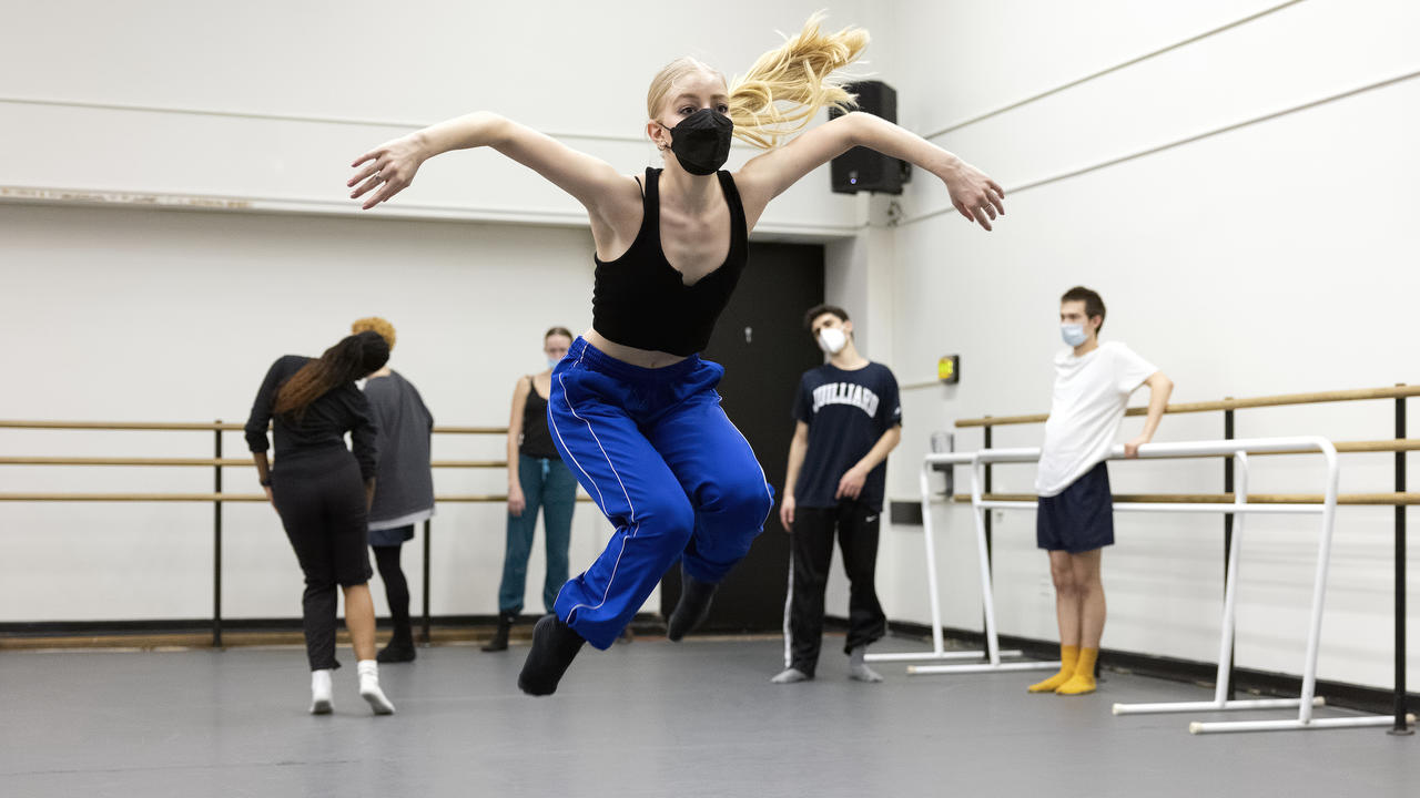 A dancer is mid-air while five others watch during a rehearsal in a rehearsal studio