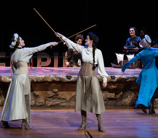 A scene from the production of 'Dido and Aeneas' featuring 2 sword fighters in the foreground and the chorus in the background