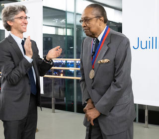 Juilliard President Damian Woetzel, left, with Clarence B. Jones, longtime Martin Luther King Jr. confidante, lawyer and speechwriter. They are standing in a dance studio, behind them are banners which show the Juilliard logo and Woetzel is applauding Jones.