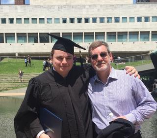 Douglas Marriner and Frank Kimbrough stand with arms around each other's shoulder in front of the Juilliard building and Marriner is dressed in his graduation cap and gown