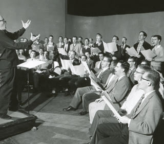 Archival (black and white) photo from a choir rehearsal of five rows of men in a semicircle. The conductor makes a passionate e