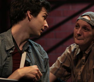 Two actors look at each other mid-scene and one actor's arm is in a cast and the other actor is wearing a backward facing baseball cap