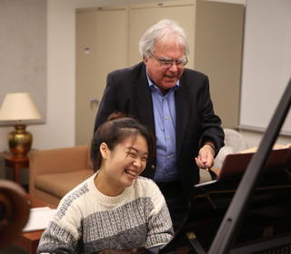 A student is playing piano and Kalichstein stands behind her, coaching