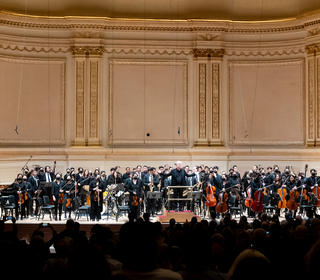 A wide-angle photo of the stage at Carnegie Hall, taken from beind the audience as the Juilliard Orchestra and Juilliard Jazz Orchestra, with David Robertson, take bows