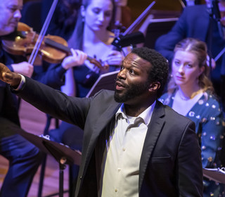 A student in performance attire is singing a solo on stage and an orchestra is visible behind him in this photo from a live performance