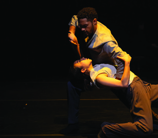Two dancers, named in the accompanying caption, perform an expressive and intimate dance on stage under dim lighting. The focus is on their intense connection and the fluidity of their movements, creating a powerful and emotional performance.