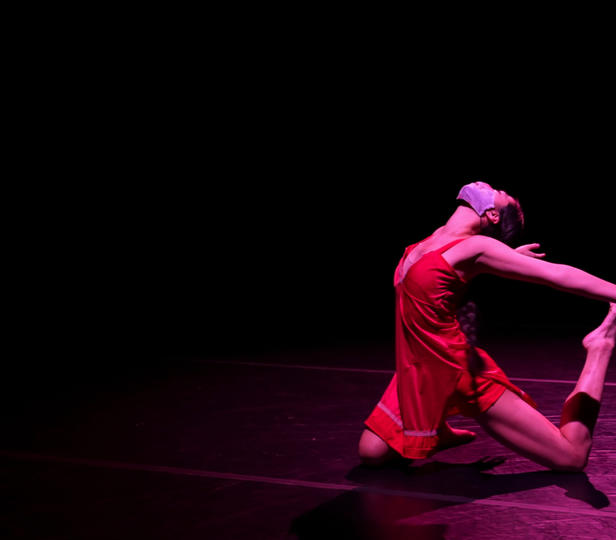 A dancer performing, costumed in a red dress in a kneeling pose with arms extended behind her
