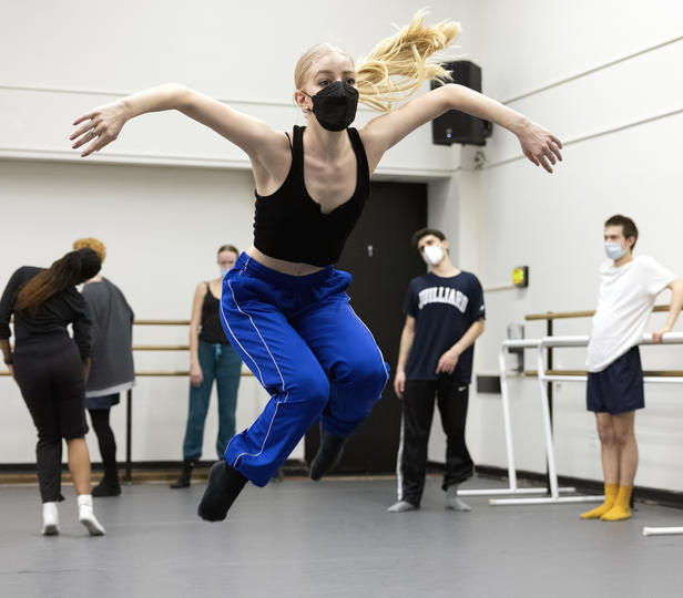 A dancer is mid-air while five others watch during a rehearsal in a rehearsal studio