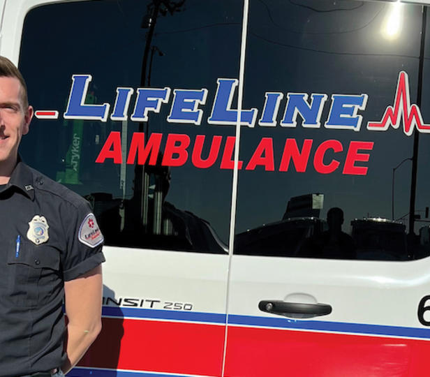 Nathan Makolandra dressed in a uniform standing beside an ambulance. On the back window of the ambulance is visible the logo "LifeLine Ambulance," which is the ambulance company Makolandra works for.