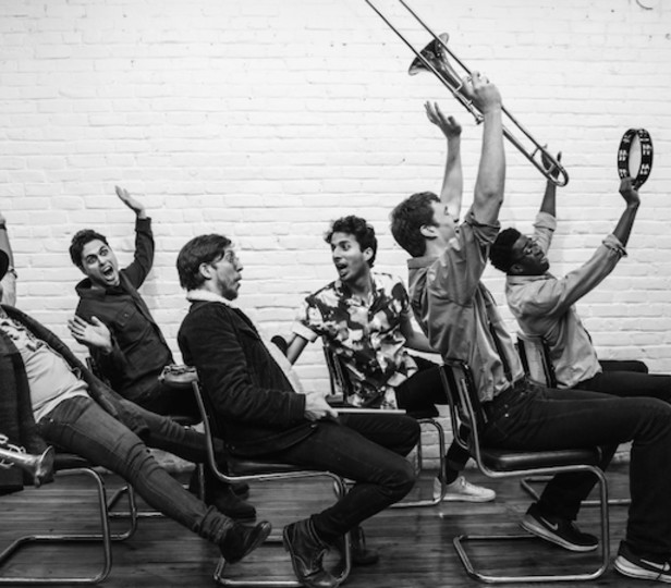 Black and white image of Sammy Miller and his group the congregation. The individuals are sitting in chairs and making a happy expression and some of them are holding their instruments (trumpets, trombone, a tambourine)