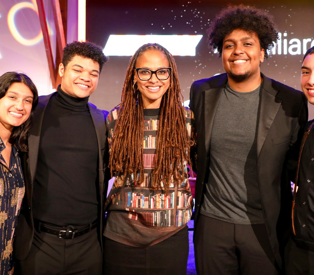 This image features a group of five individuals posing together for a photo. They all appear to be cheerful and are standing in front of a backdrop that includes the word "Juilliard" in large, stylized letters, suggesting that they are at a special event.