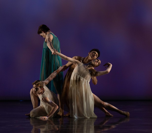 ​​​​​​​Four dancers on stage. One in the foreground is crouched on the floor, propping themselves up with one arm while looking attentively to the side. Behind them, a dancer in a flowing white dress is caught in mid-movement, arms elegantly extended and one leg bent at the knee in a pose that suggests fluid motion. The dancer leans back against another dancer who is standing upright, supporting them with one arm and reaching forward with the other.