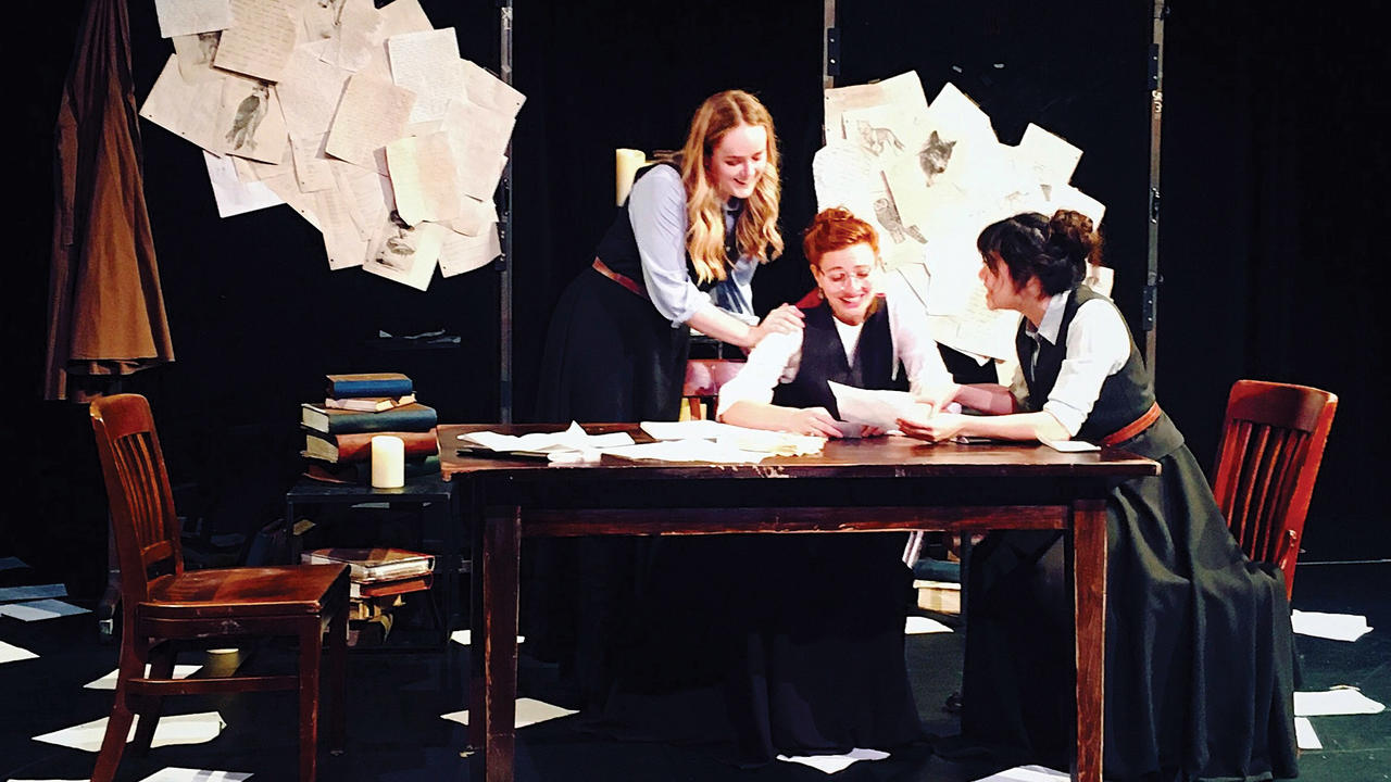 A scene from 'Brontë.' The set features three of the cast seated at a wooden table. The room and tabletop are littered with papers.