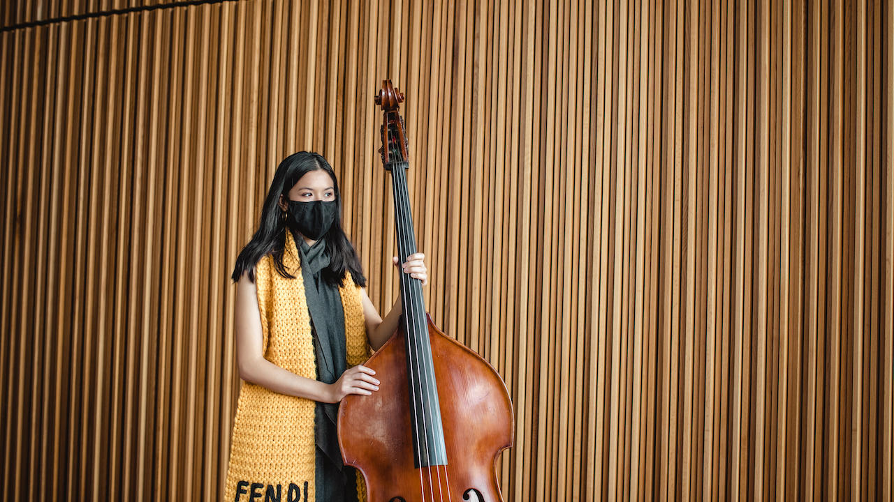 A bass player, dressed in a distinctive yellow Fendi scarf, holds her bass in front of a wood-paneled wall in the Peter Jay Sharp Theater