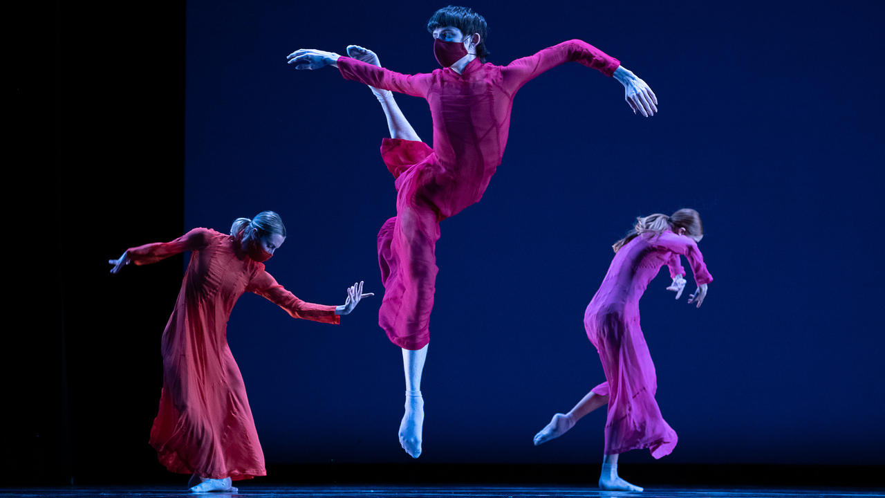 Three dancers costumed in red in the midst of performance. One dancer in the middle is suspended in the air.