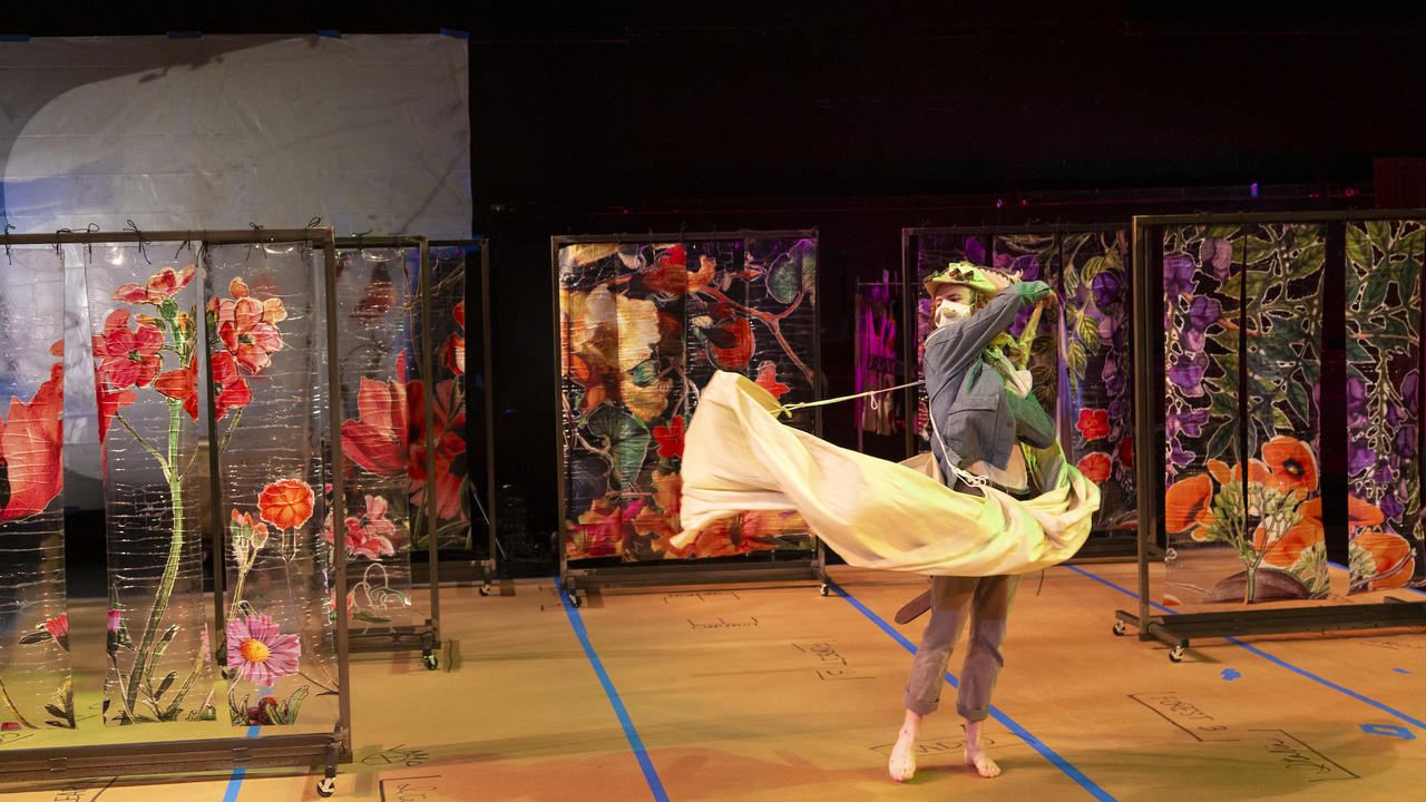 A actor, wearing a face mask, is on stage and the set consists of multi-colored paintings of flowers