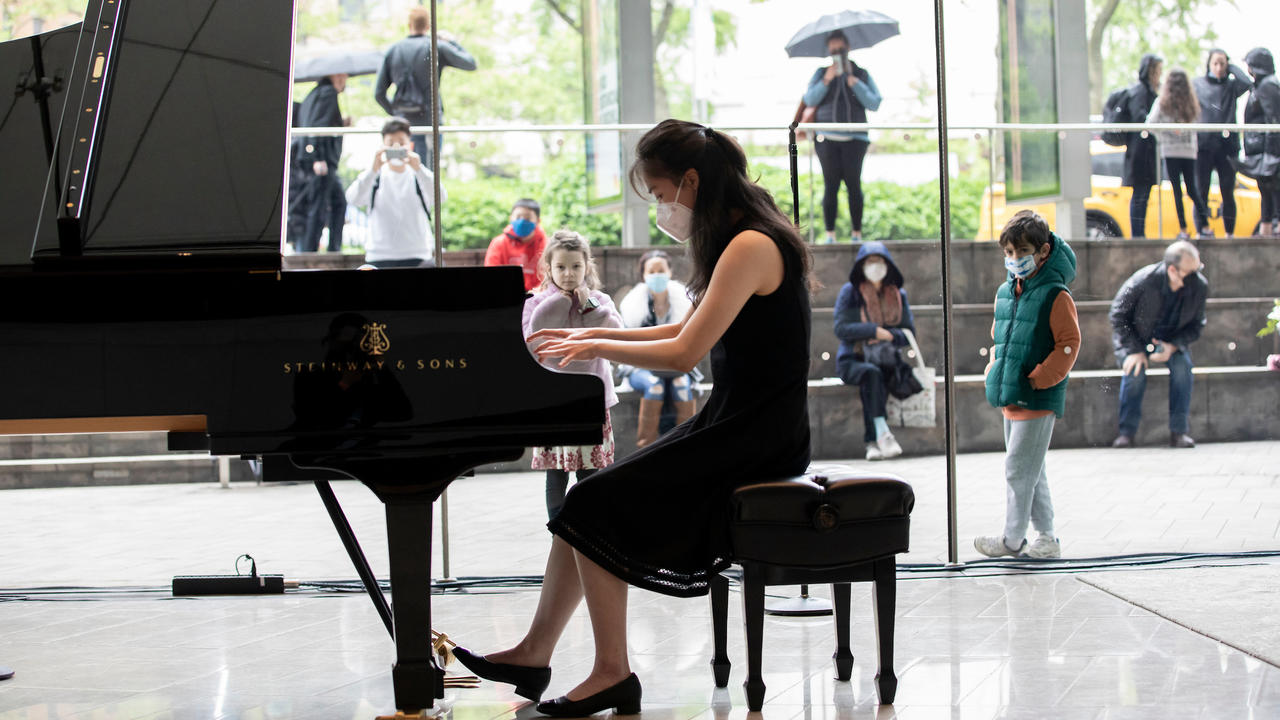 Katherine Wang is playing piano in the lobby of Alice Tully Hall and onlookers are visible through the glass