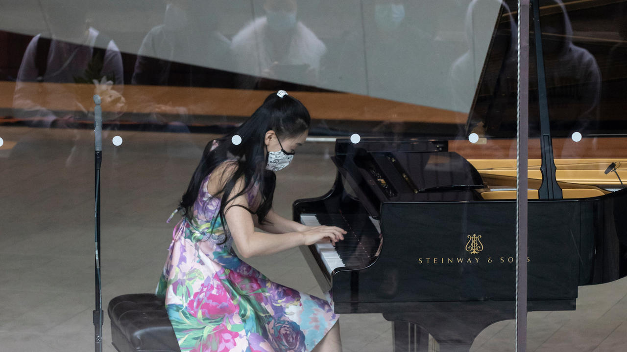 Harmony Zhu is playing piano in the lobby of Alice Tully Hall and the reflections of onlookers (passersby) are visible in the glass pane