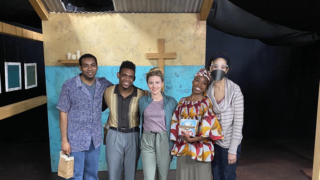 Jade King Carroll poses with four cast members from her show in front of a piece of the set that looks like a small church. A wooden cross is seen hanging on the wall behind them and a small wooden shelf has several votive candles on it.