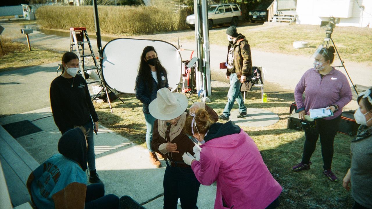 Outside on a sunny day, some crew members and cast members are seen working together and there is some filming equipment visible around them including for lighting and microphones and a ladder