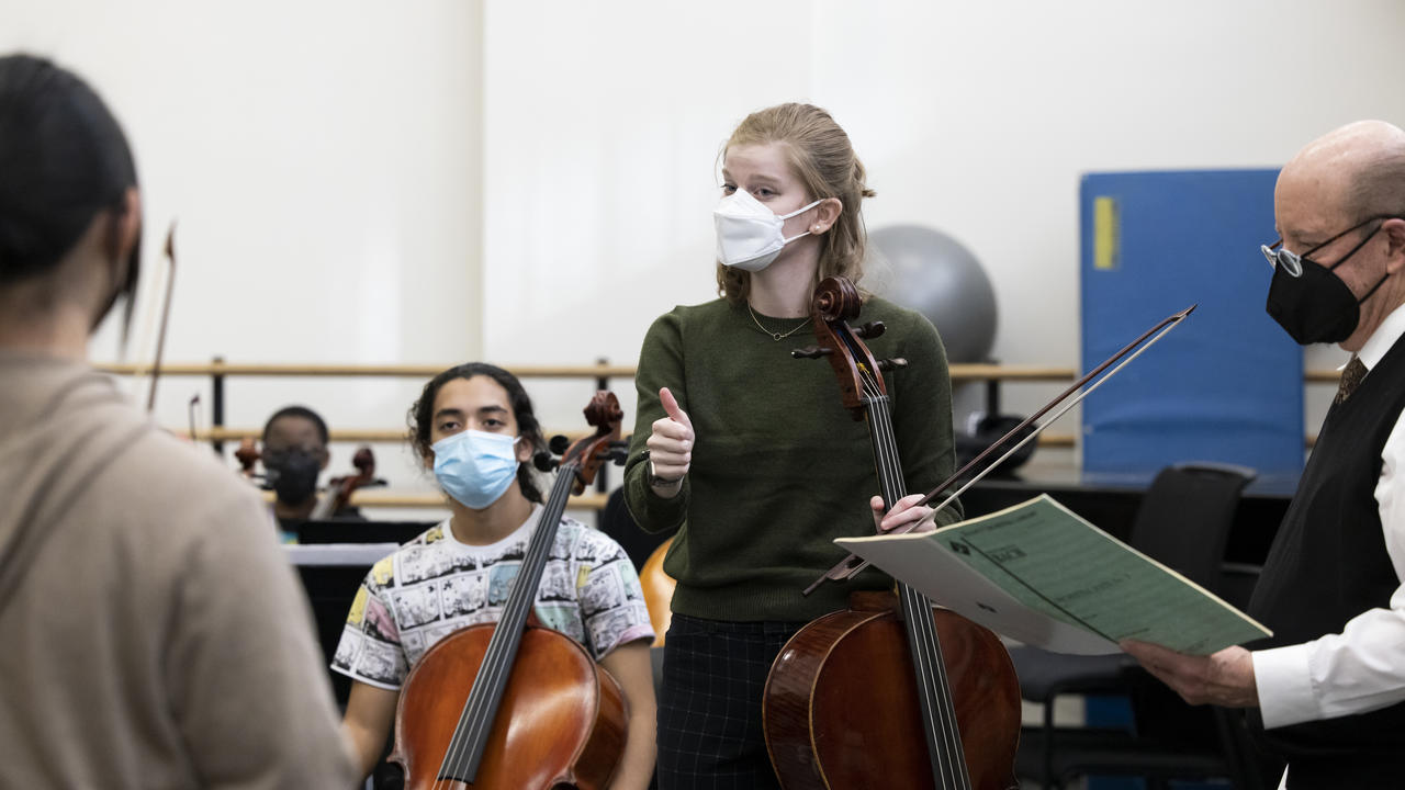 Adrienne Hyde is standing, holding her cello, and addressing students in the rehearsal room