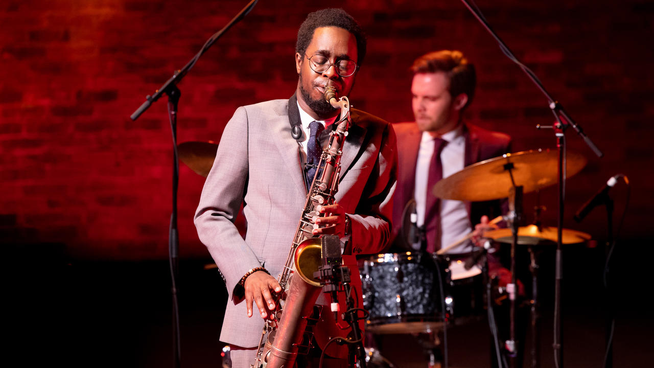 A jazz musician is playing saxophone, his eyes closed, in reverie and a percussionist is visible behind him. They are smartly dressed. The background is a brick wall.