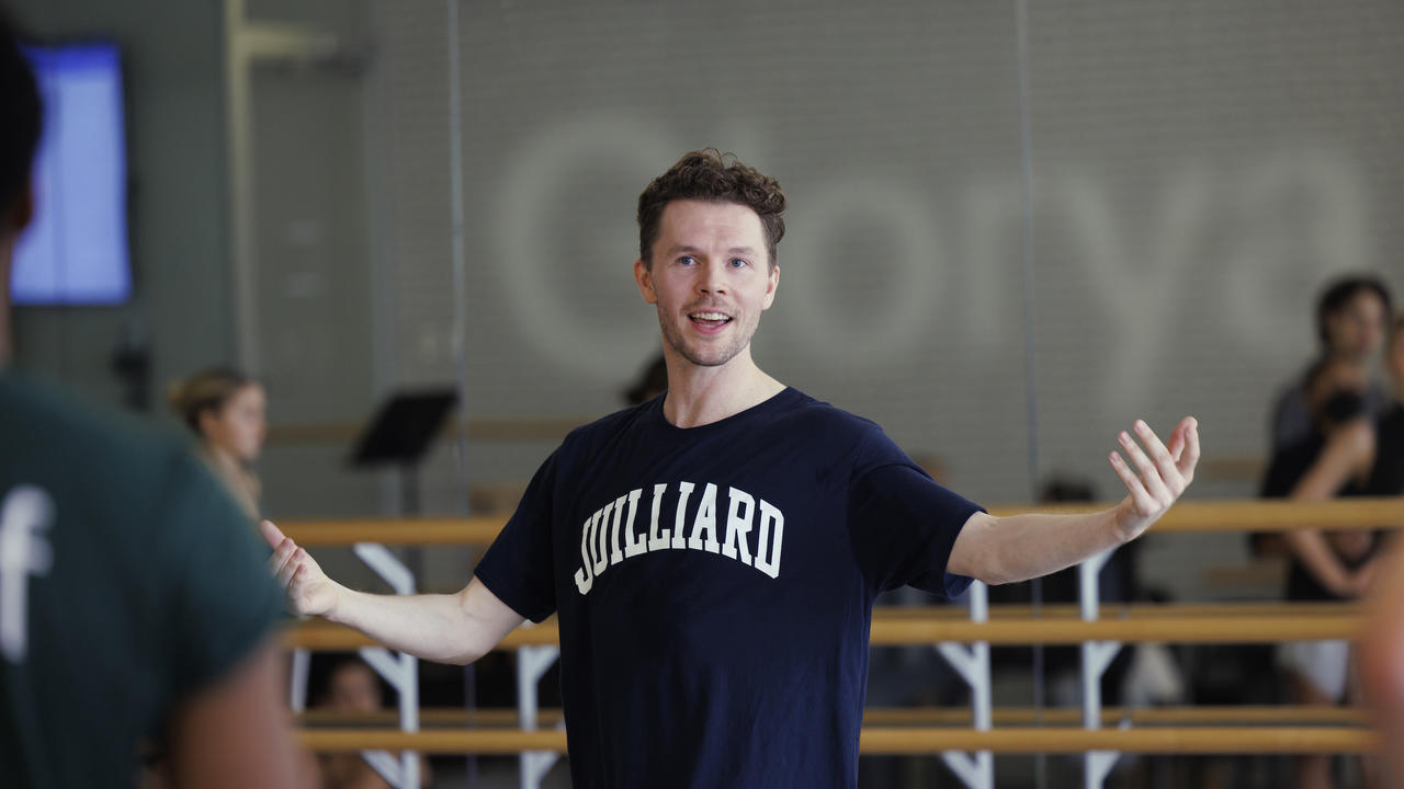 Spenser Theberge wearing a blue Juilliard tee standing in a dance studio, speaking to a group of dancers who are visible in the mirror behind him, and making a gesture with arms extended wide