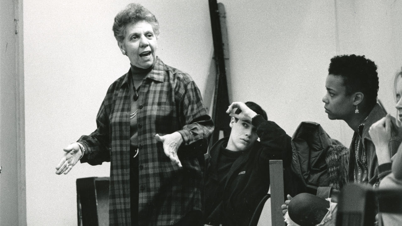 Eve Shapiro in a black and white (archival) photograph makes an emphatic expression while directing or teaching and students watch with intent