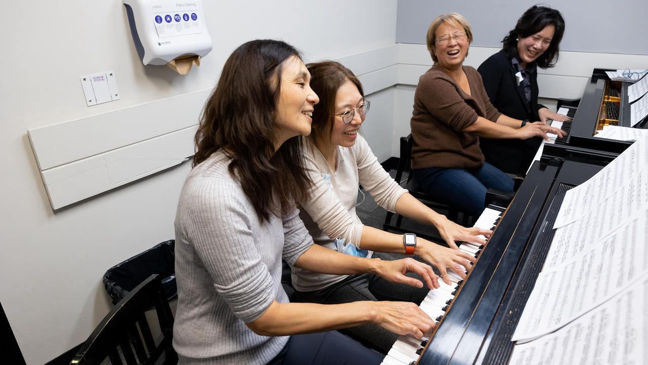 Four piano players in a practice studio sit at each of two pianos, playing four-hands arrangement. They are smiling and laughing.