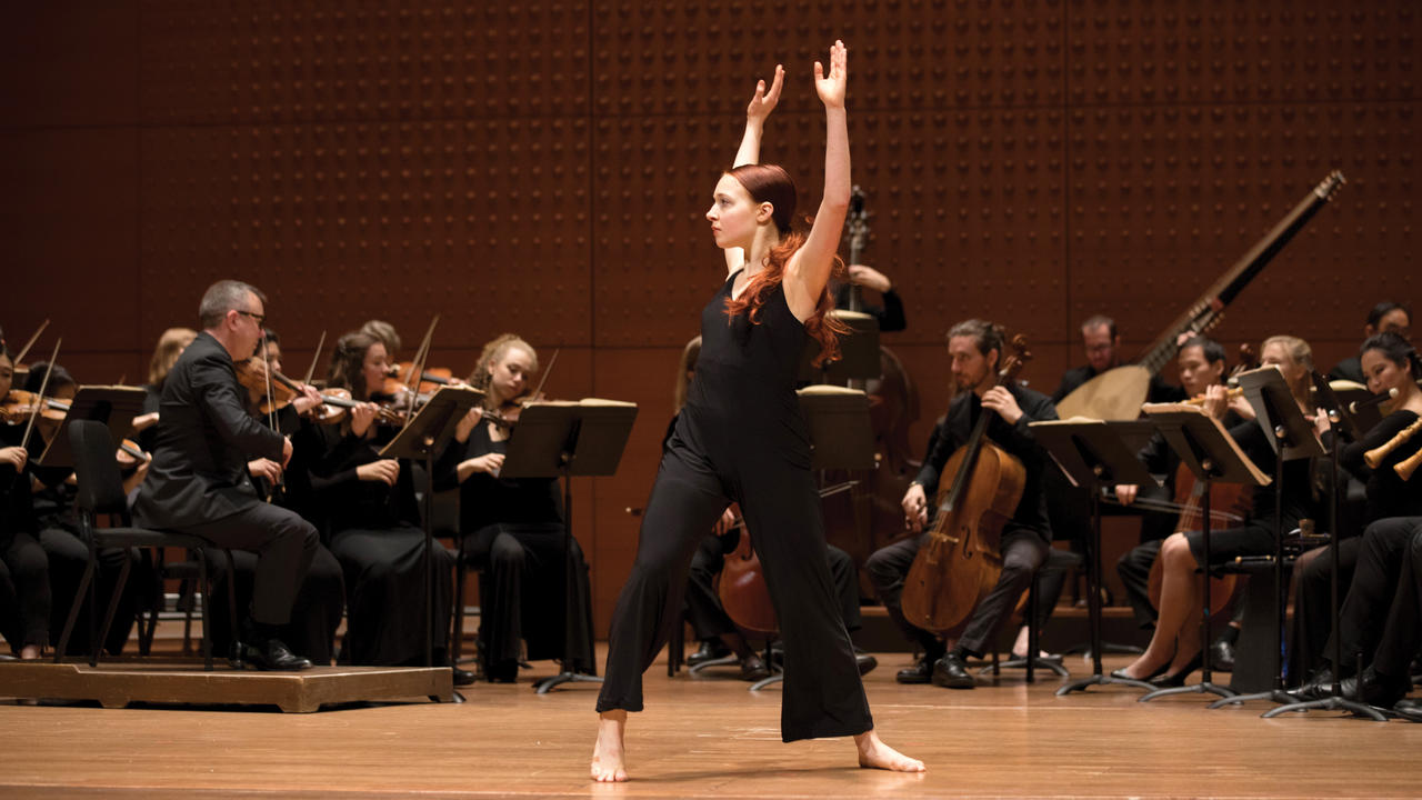 A dancer on stage performing and behind her is an orchestra. The picture communicates the idea of interdisciplinary performance.