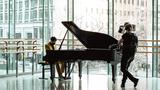 A pianist, dressed in Fendi, playing a grand piano in a dance studio with the camera person included to communicate a sense that this is behind-the-scenes