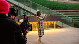 A trumpet player, dressed in a distinctive knee-length Fendi coat, plays outdoors in Lincoln Center Plaza and a camera person and their equipment is in the foreground