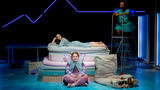 Three actors, one dressed like a fairy, are in a bedroom and one actor is lying on a pile of mattresses