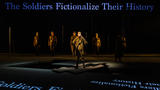 A group of actors on stage, mid-performance, are dressed like soldiers. The words "The Soldiers Fictionalize Their History" are being projected onto the stage and scenery.